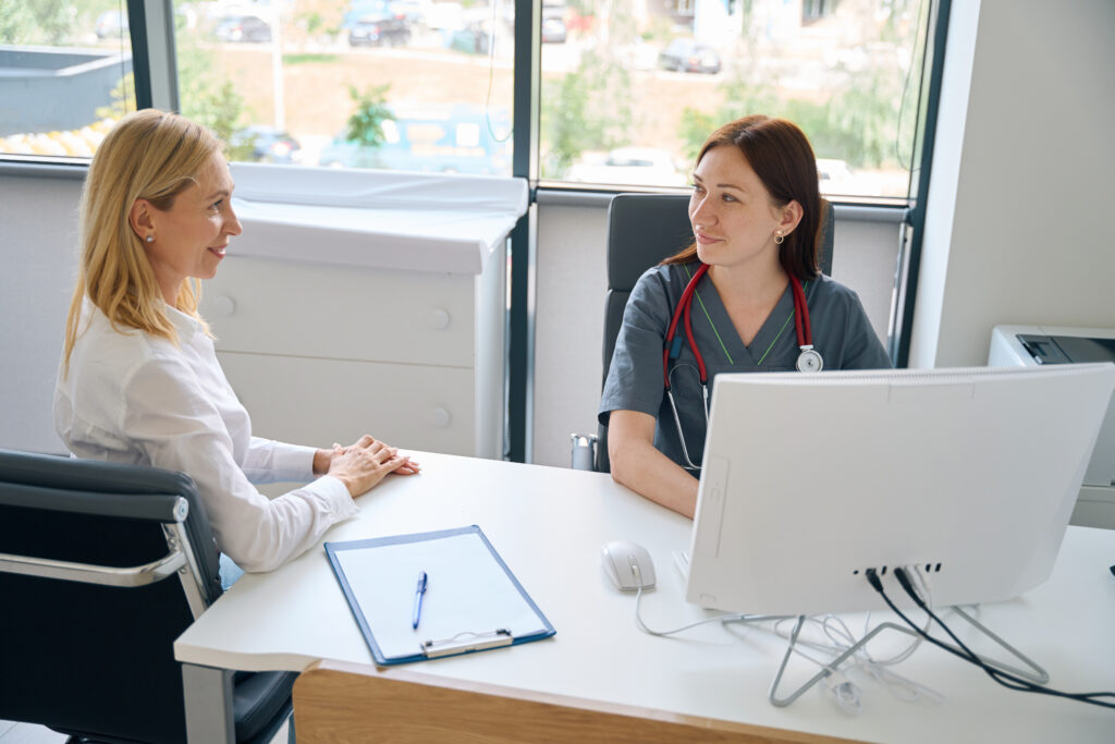 General practitioner seated at desk in front of PC listening to female client during primary consultation